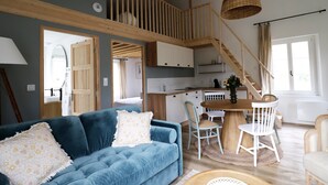 A modern living room with a blue sofa, white chairs, a wooden staircase, and a kitchen area. - Le Domaine Saint Ange (Friville-Escarbotin)