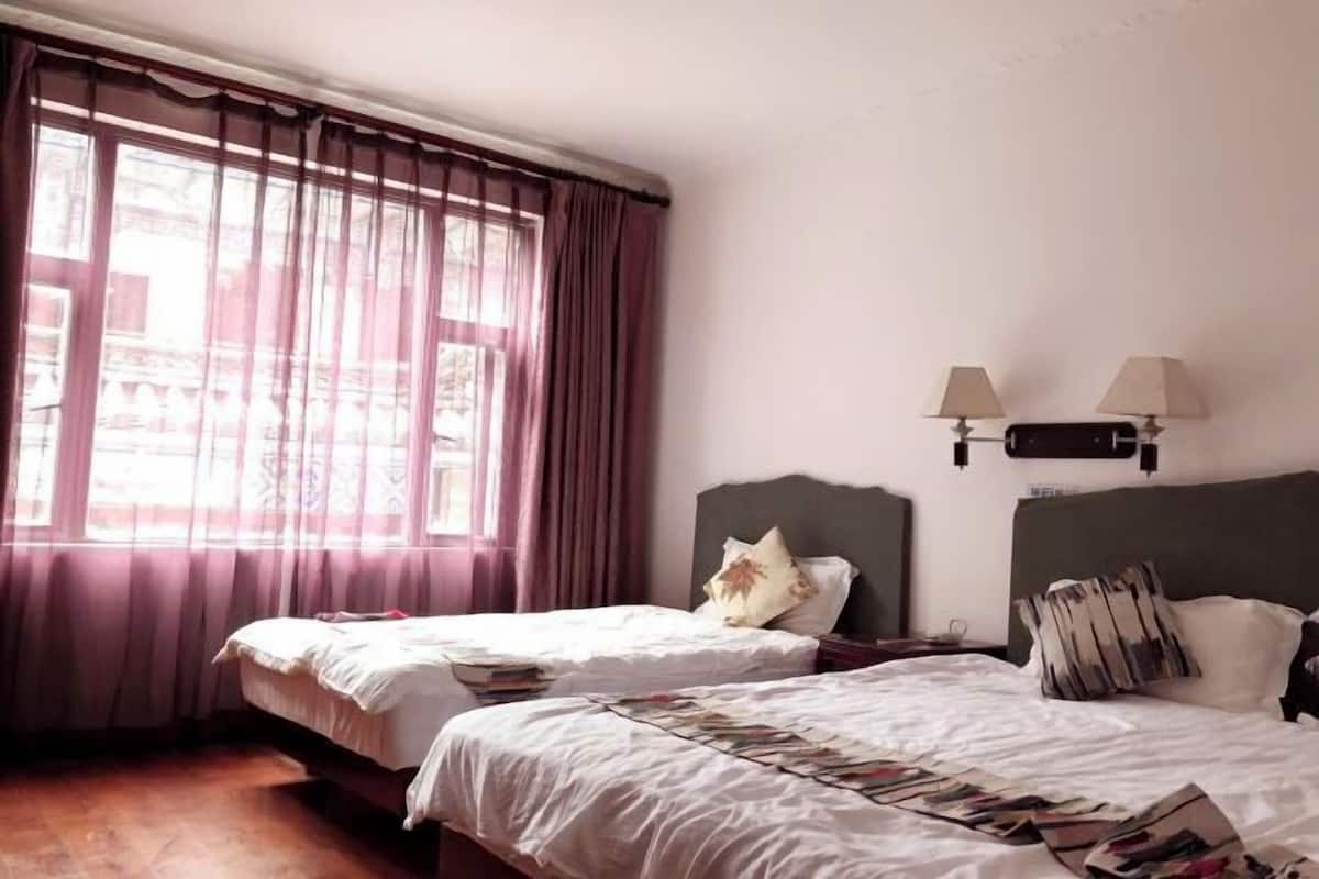 A hotel room with two beds, a window with curtains, a nightstand with lamps, and a wall-mounted light fixture.