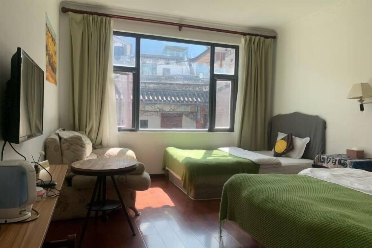 A hotel room with a large window, a desk with a computer, a chair, two beds, and a small table.