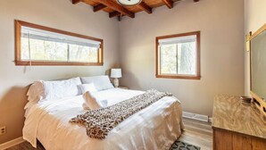 Un dormitorio con una cama grande, dos ventanas, techo de madera y una mesita de noche con lámpara.