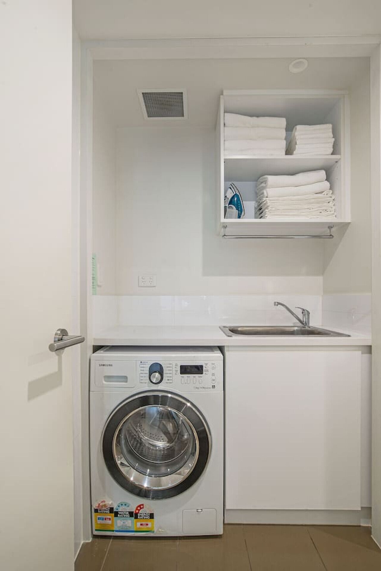 Un espace buanderie compact comprenant un lave-linge à chargement frontal, un évier et une étagère au-dessus pour ranger les serviettes pliées.