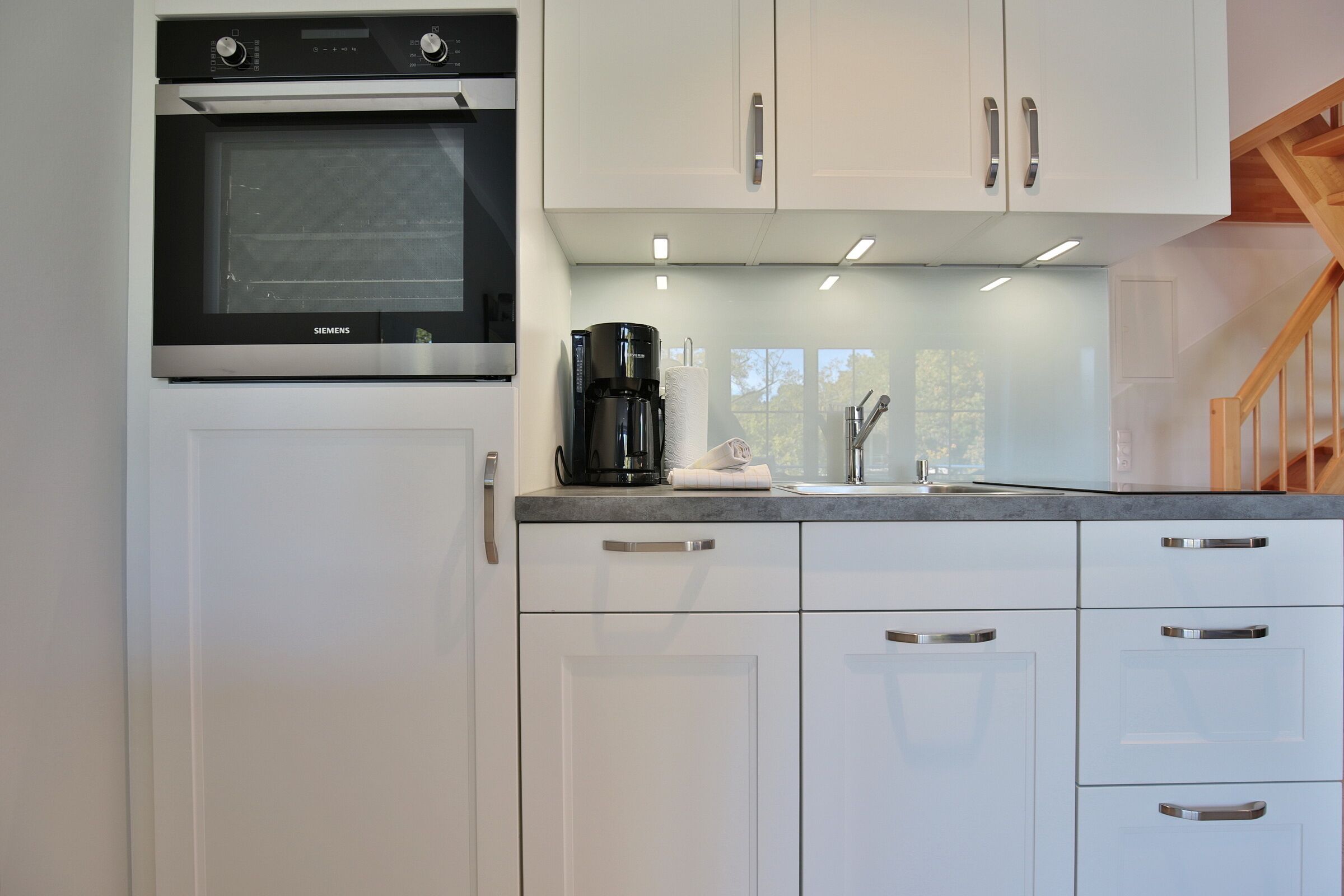 Una cocina moderna con armarios blancos, horno empotrado y cafetera.