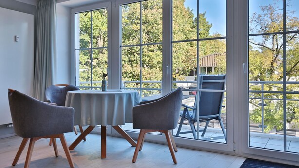 A room with a large window, a table with chairs, and a view of trees and a balcony. - Haus Schäfer Appartement 8 (Timmendorfer Strand)