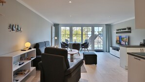 A modern living room with a sofa, a coffee table, and a TV. There is a dining area with chairs and a table. The room has large windows and a view of the outdoors. - Haus Schäfer Appartement 8 (Timmendorfer Strand)