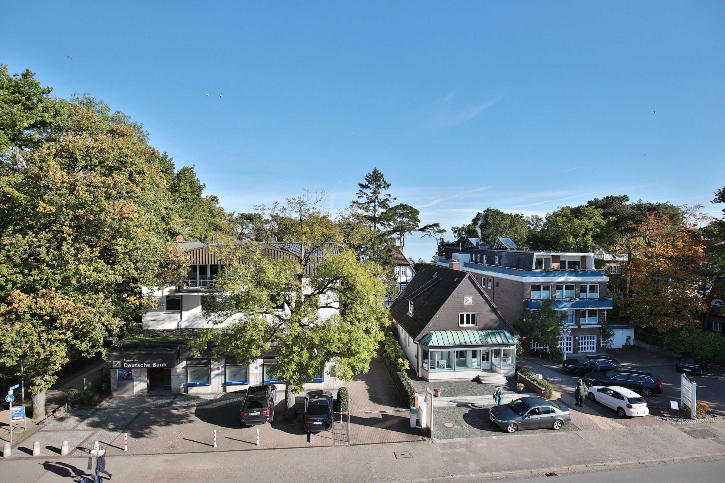 A multi-story building with a green roof and a sign for Deutsche Bank. There are parked cars and a pedestrian walkway.