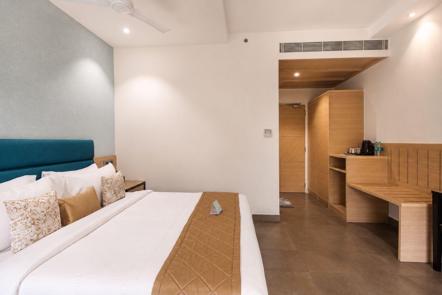A modern hotel room with a large bed, a wooden bench, and a kitchenette area.