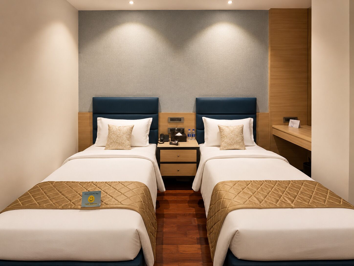A hotel room with two beds, a wooden floor, a nightstand with a phone and a lamp, and a wall-mounted shelf.