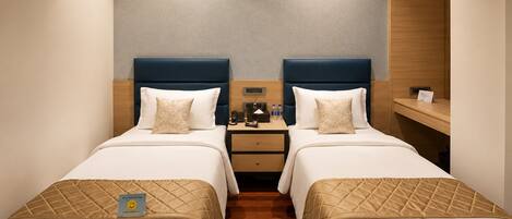 A hotel room with two beds, a wooden floor, a nightstand with a phone and a lamp, and a wall-mounted shelf.