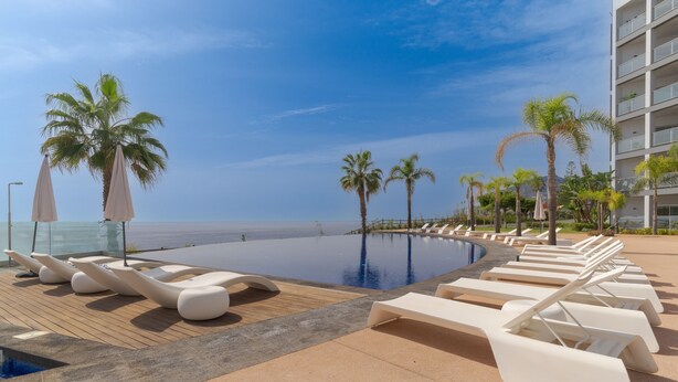 A poolside area with lounge chairs and umbrellas, palm trees, and a view of the ocean. - Madeira Palace Ocean Sounds (Funchal)