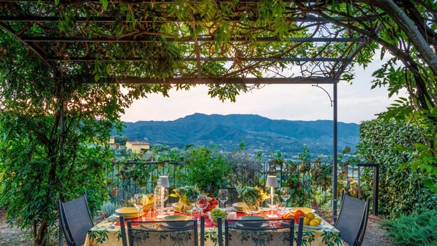 An outdoor dining area with a table set for two, surrounded by greenery and a mountain view. - Villa Debby in Lucca (Lucca)