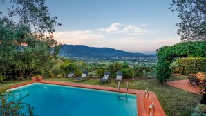 A swimming pool with lounge chairs and a table, surrounded by greenery and a view of distant mountains. - Villa Debby in Lucca (Lucca)