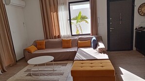 A living room with a sofa, ottoman, and a small table. There is a window with curtains, a door, and a clock on the wall. - Charme de L'atypique (Nabeul‎)