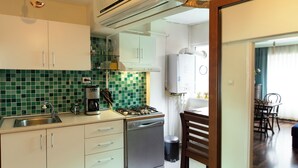 Une cuisine équipée d’une cuisinière à gaz, d’un évier et de meubles blancs. On aperçoit également, à l’arrière-plan, une machine à café, une poubelle et un coin repas.