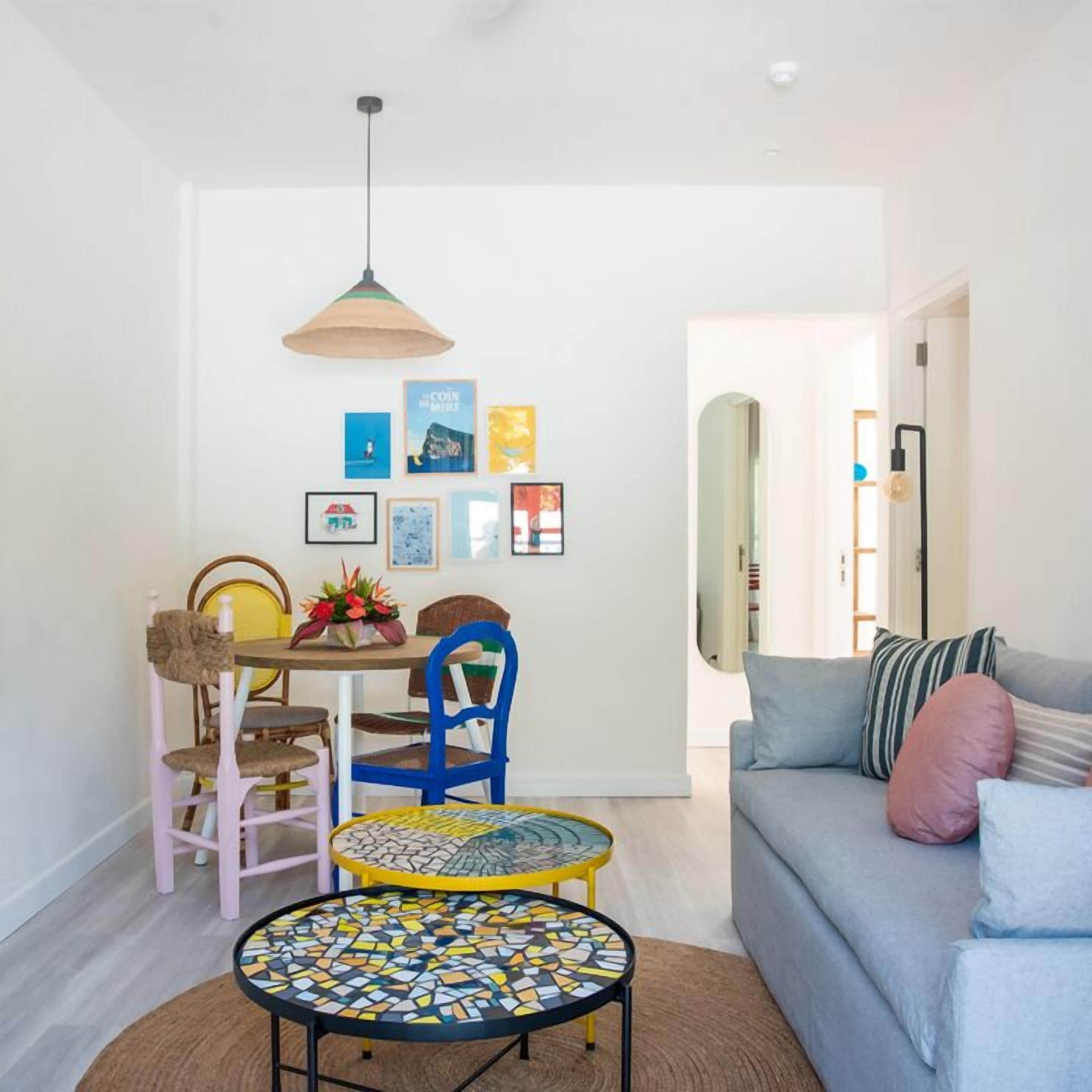 A modern living room with a gray sofa, a round coffee table with mosaic tiles, a dining area with a wooden table and chairs, and a hanging pendant light.