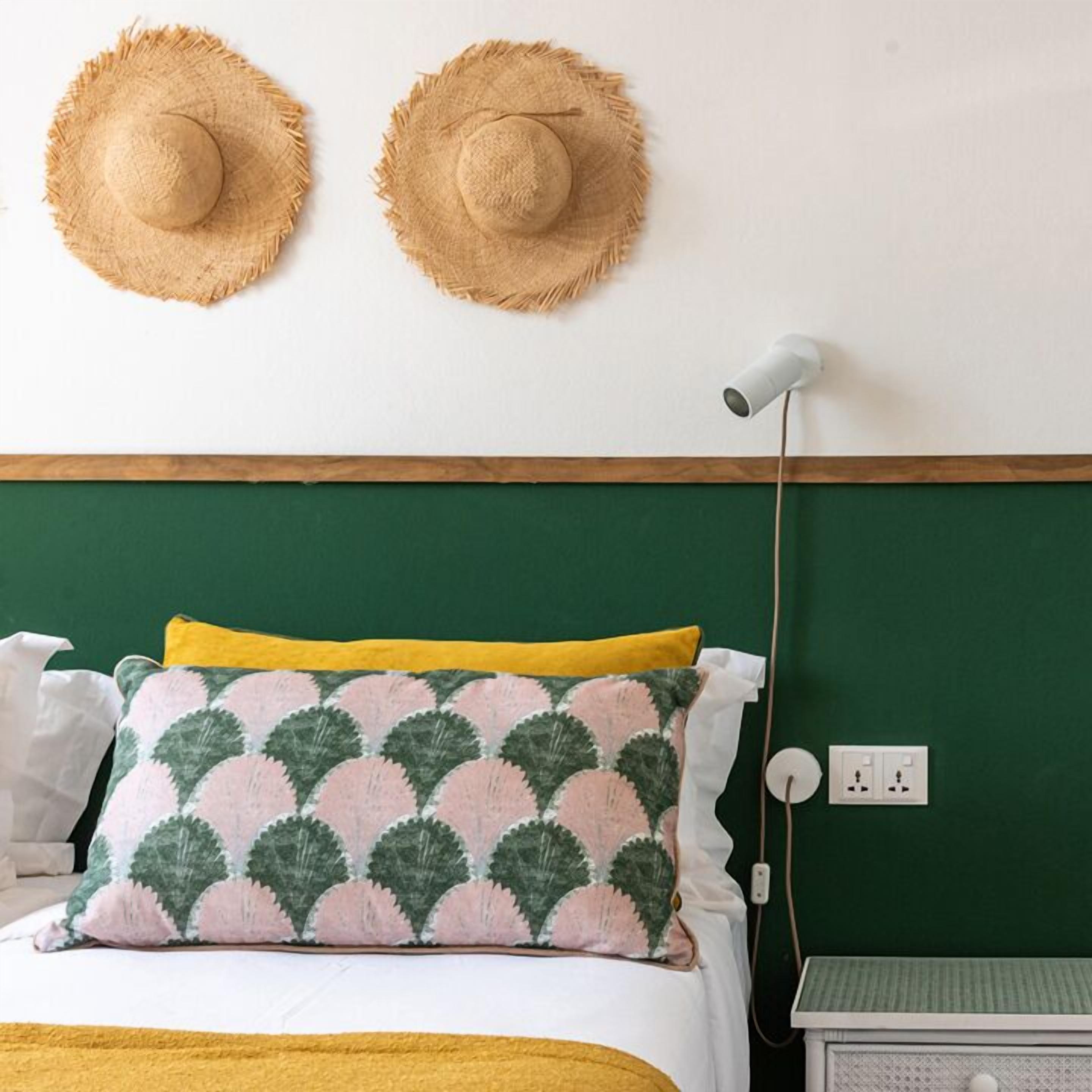 A bedroom with a green headboard, two straw hats on the wall, a patterned pillow, and a bedside table with a lamp.