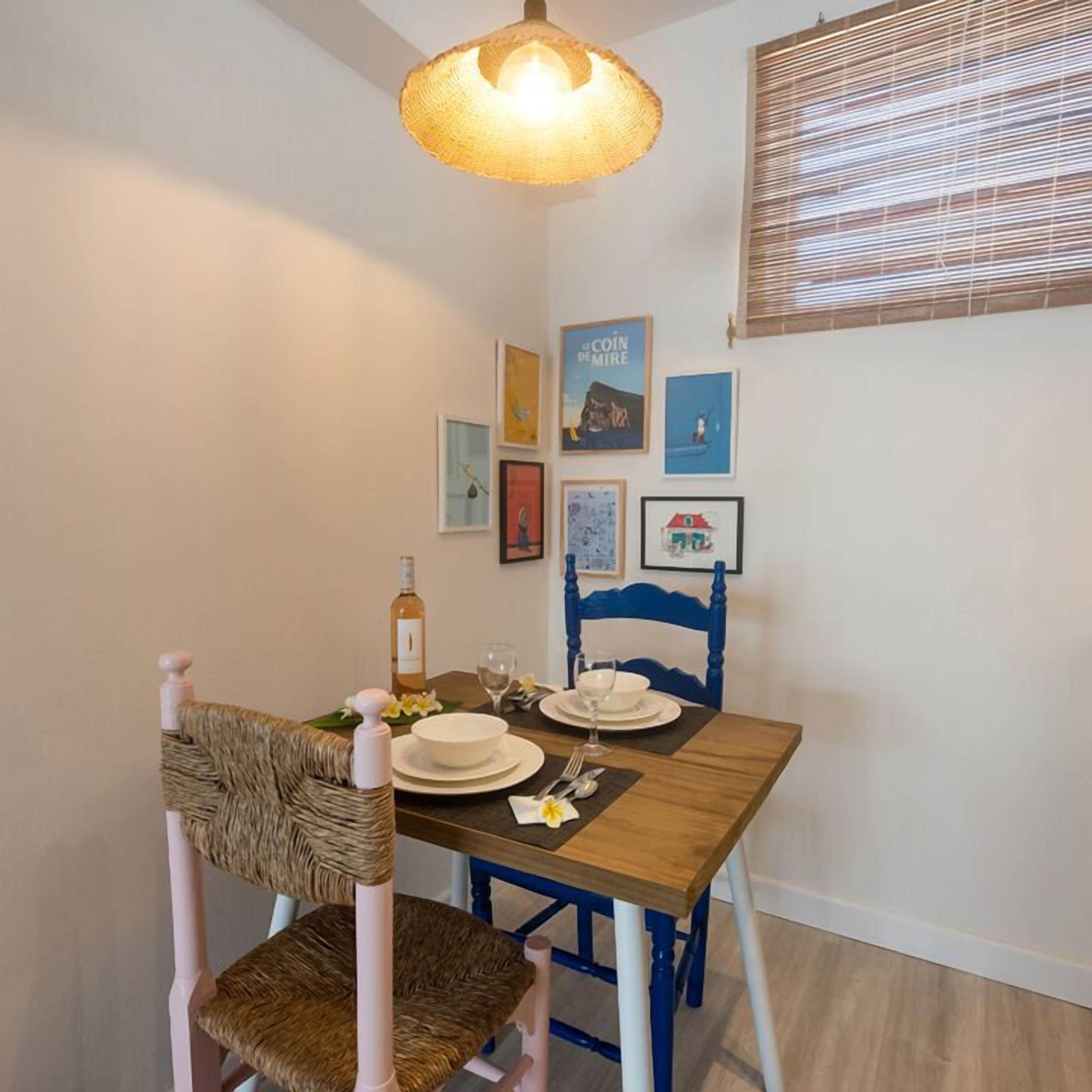 A dining area with a table set for two, a wicker chair, a blue chair, and a hanging light fixture.