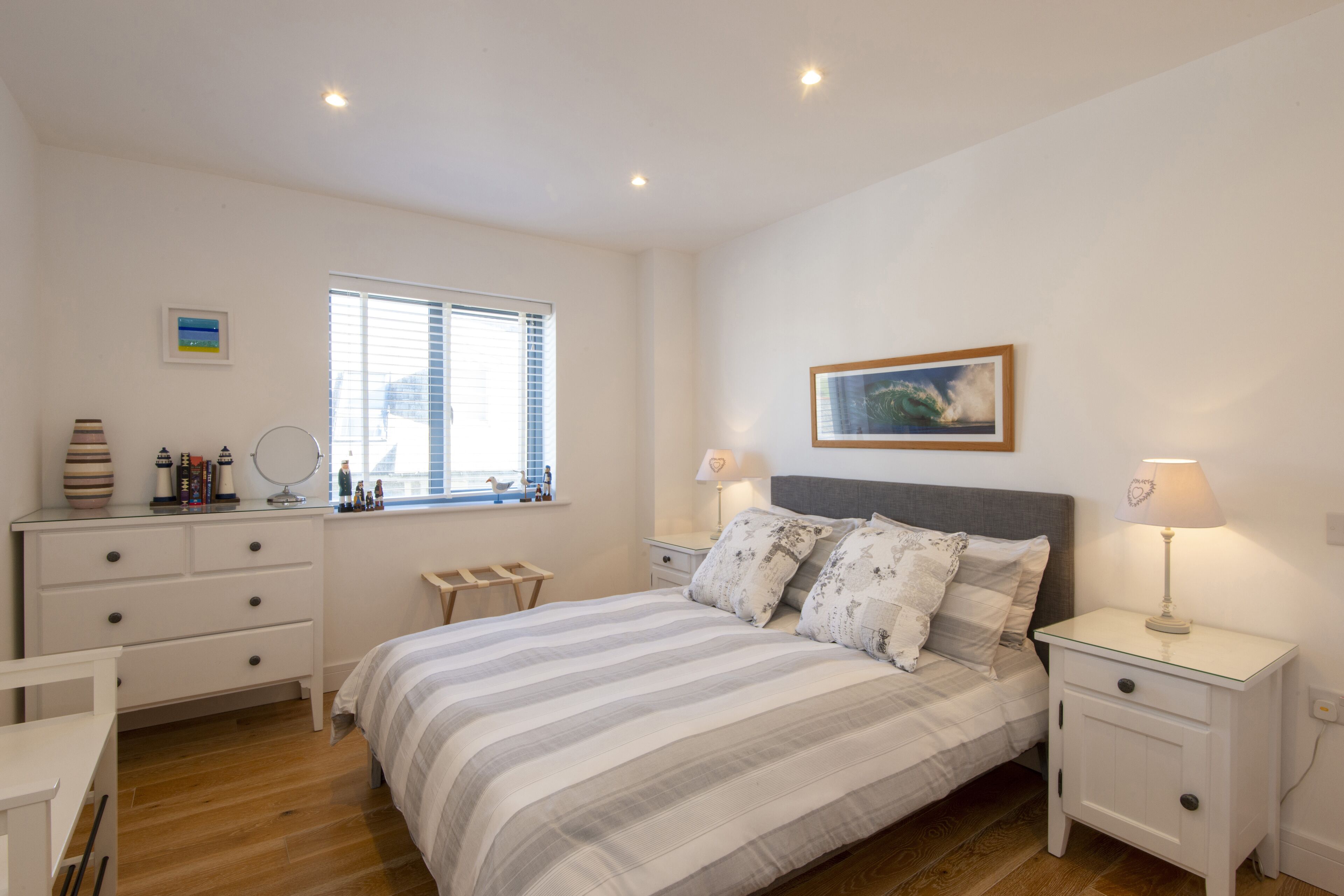 Un dormitorio con cama, mesitas de noche, cómoda, ventana con persianas e imagen enmarcada en la pared.