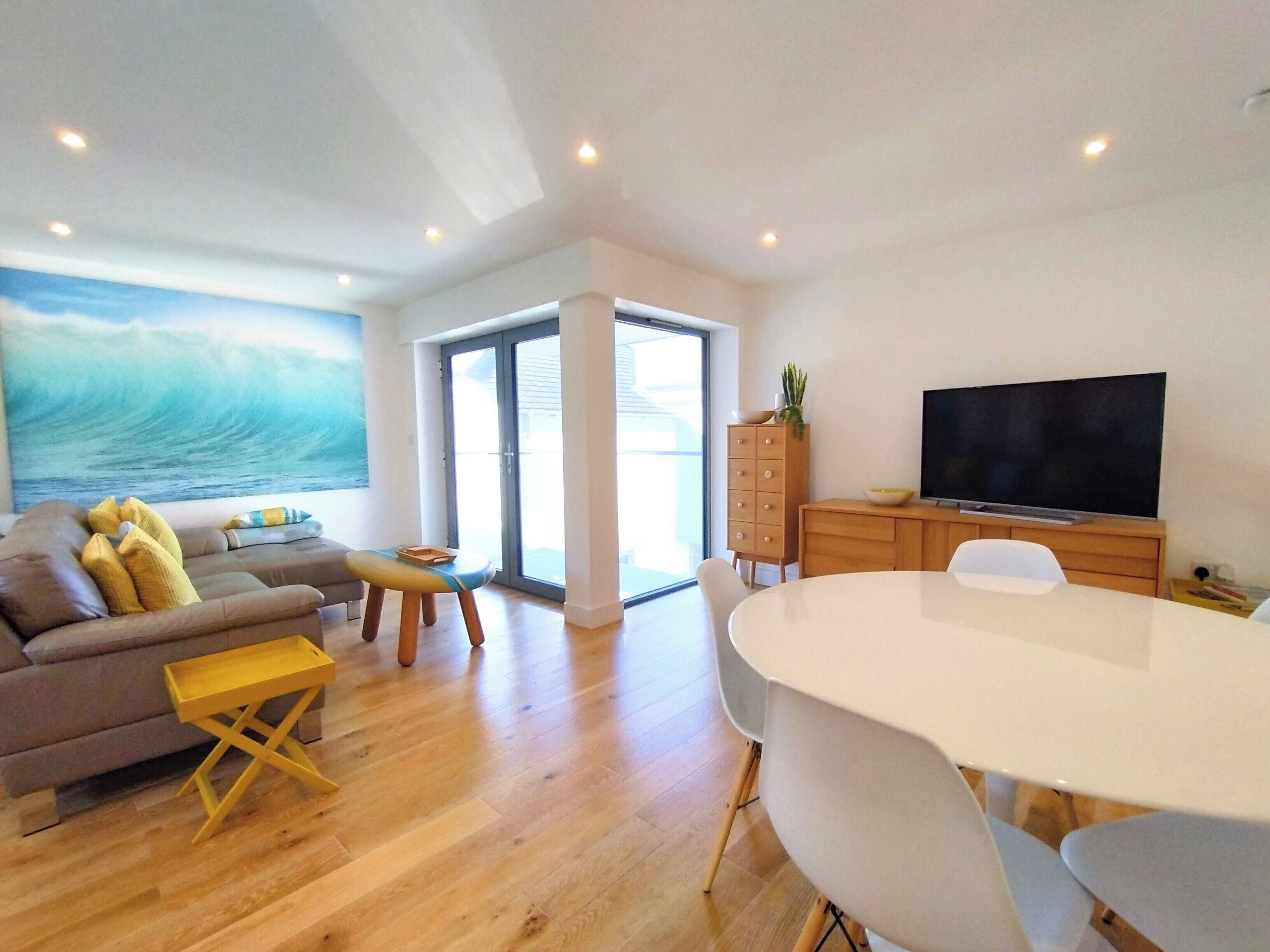 A modern living room with a sofa, a small table, a dining table with chairs, a television, and a large window with a view of the ocean.