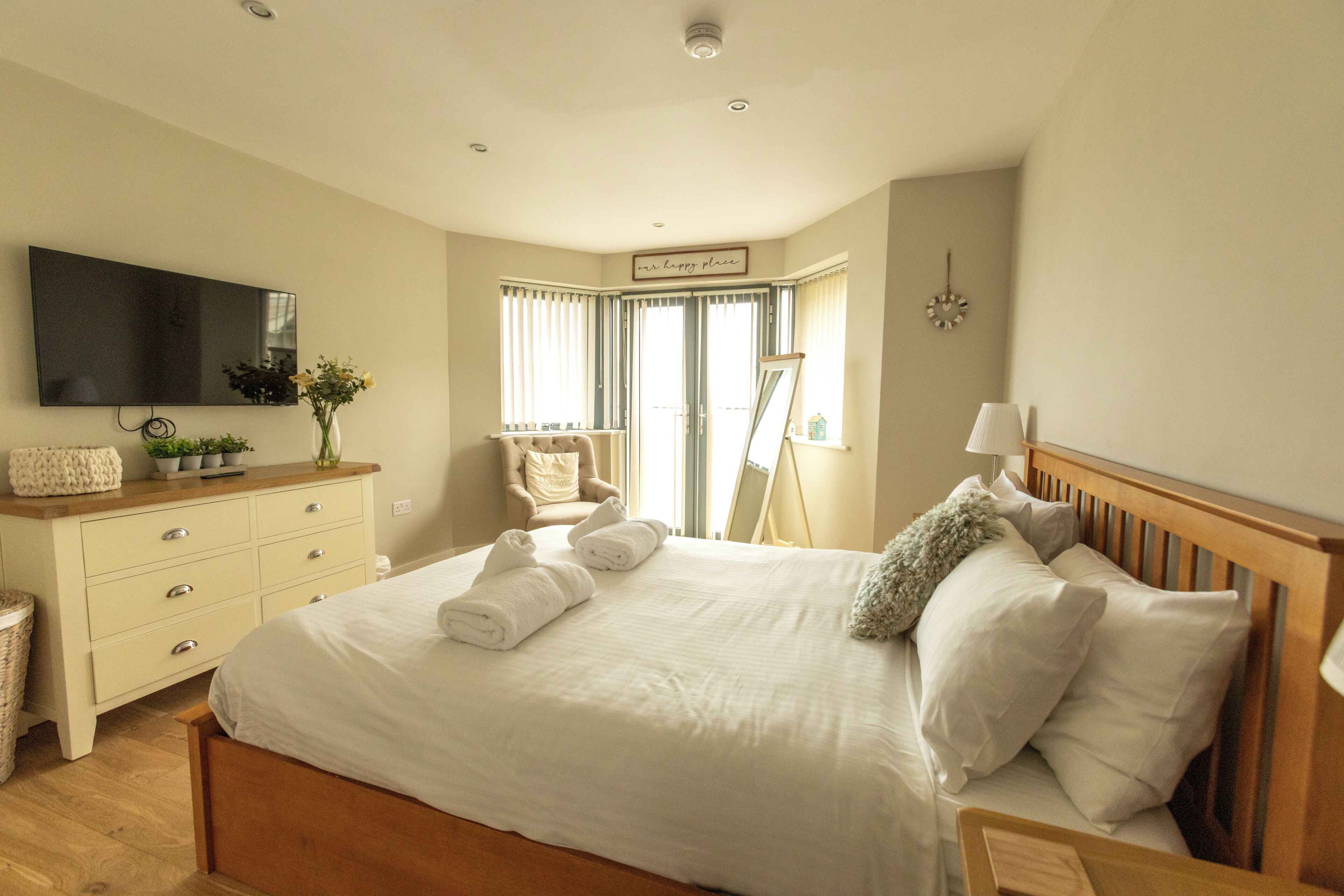 Une chambre à coucher comprenant un lit, une commode, un téléviseur, une chaise et une fenêtre avec des rideaux.