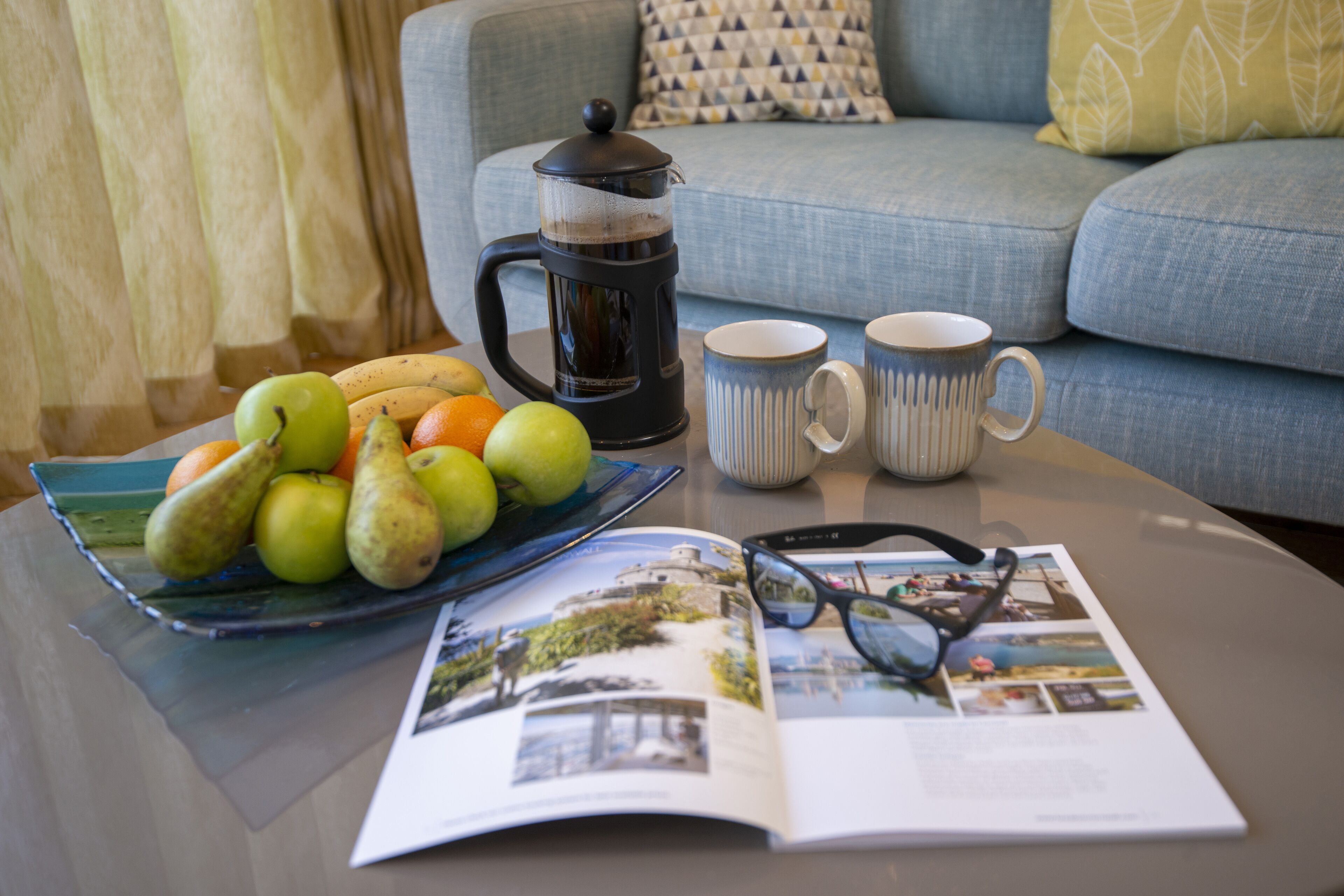 A table with a coffee maker, two mugs, a plate of fruit, and a magazine.