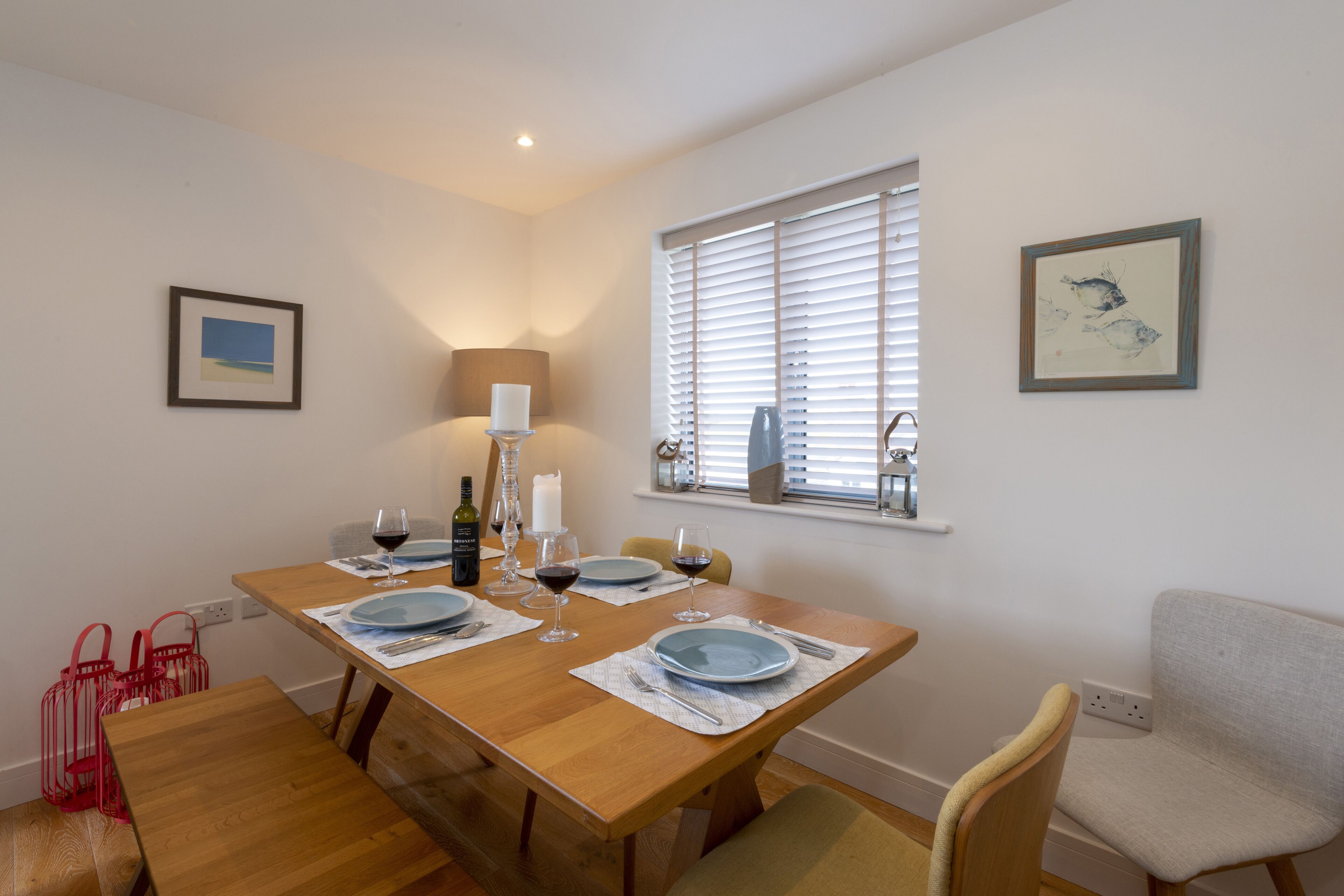 A dining area with a wooden table set for four, a window with blinds, and a lamp.