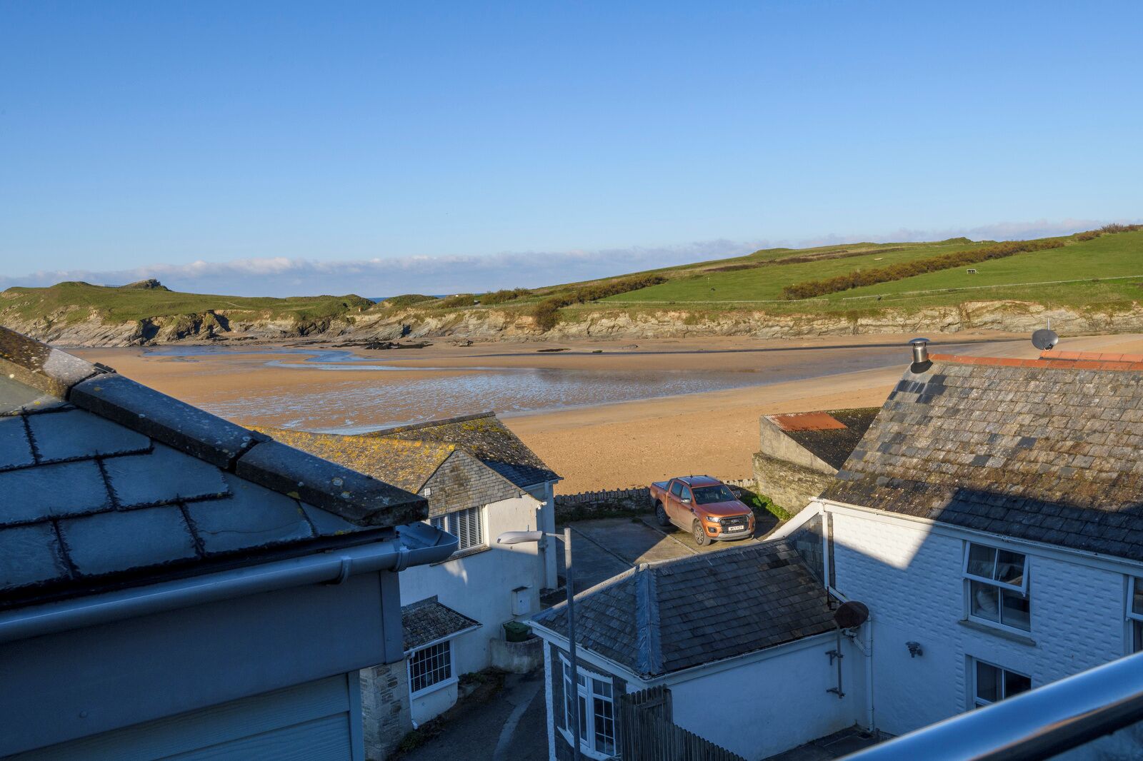 A coastal view with a sandy beach, a parked car, and white buildings with slate roofs.