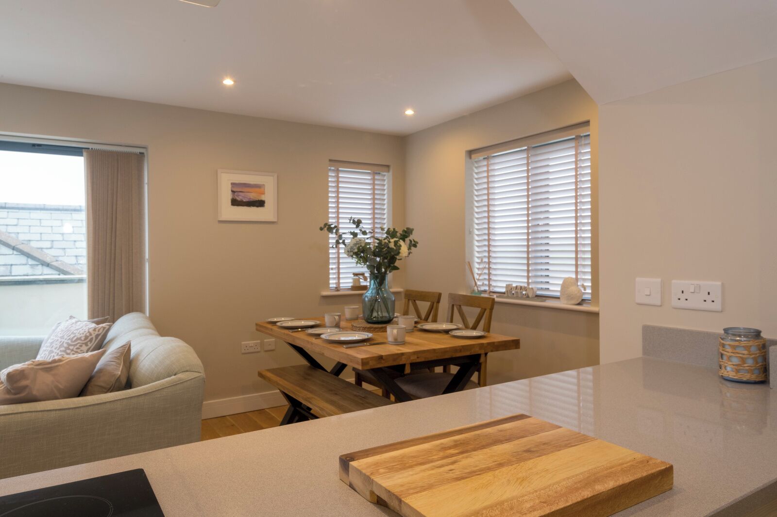 Una cocina moderna con una mesa de madera lista para comer, una encimera con tabla de cortar y un florero con flores en el alféizar de la ventana.