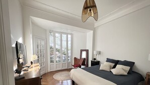 Een slaapkamer met een groot bed, een stoel, een bureau en een spiegel. Er zijn grote ramen en een deur die naar een buitenruimte leidt.