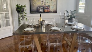 Een eetgedeelte met een grote tafel voor zes personen, transparante stoelen en een fles champagne. Aan de muur hangt een groot schilderij, en ernaast staat een potplant.