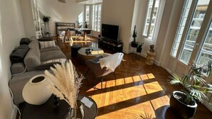 Een ruime woonkamer met een grote bank, een salontafel en een televisie. Er zijn ramen die veel natuurlijk licht binnenlaten en uitzicht bieden op buiten.