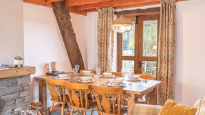 Un coin repas chaleureux avec une table en bois dressée pour quatre personnes, entourée de chaises en bois. On peut apercevoir un mur en pierre et un plafond avec une poutre en bois.