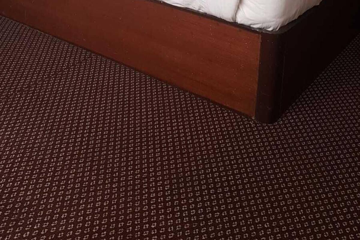 A hotel room with a bed, a headboard, a wall-mounted air conditioner, and a patterned carpet.