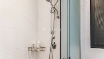 A modern shower with a handheld showerhead and a wall-mounted faucet. There is a small shelf with two bottles of shampoo and conditioner.