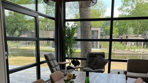 A dining area with a large wooden table, chairs, and a view of a garden through large windows. - Ferienhaus Holland-cube Direkt am Veluvemeer in Bad Hoophuizen (Hulshorst)