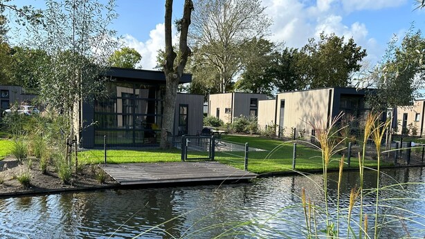 A modern building with a wooden deck over a water body, surrounded by greenery and trees. - Ferienhaus Holland-cube Direkt am Veluvemeer in Bad Hoophuizen (Hulshorst)