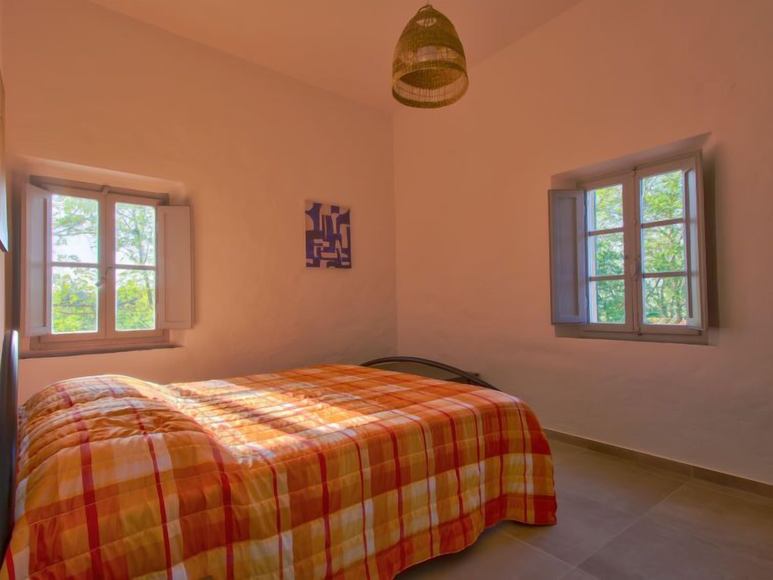 A bedroom with a bed, two windows, a hanging light fixture, and a wall-mounted artwork.