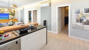 Une cuisine moderne dotée d’un grand îlot central, un coin repas avec une table en verre et un salon orné d’un tableau représentant une plage.