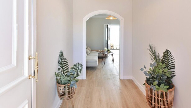 Un pasillo con piso de madera, paredes blancas y una puerta a la izquierda. Hay dos cestas de mimbre con plantas a cada lado del pasillo.