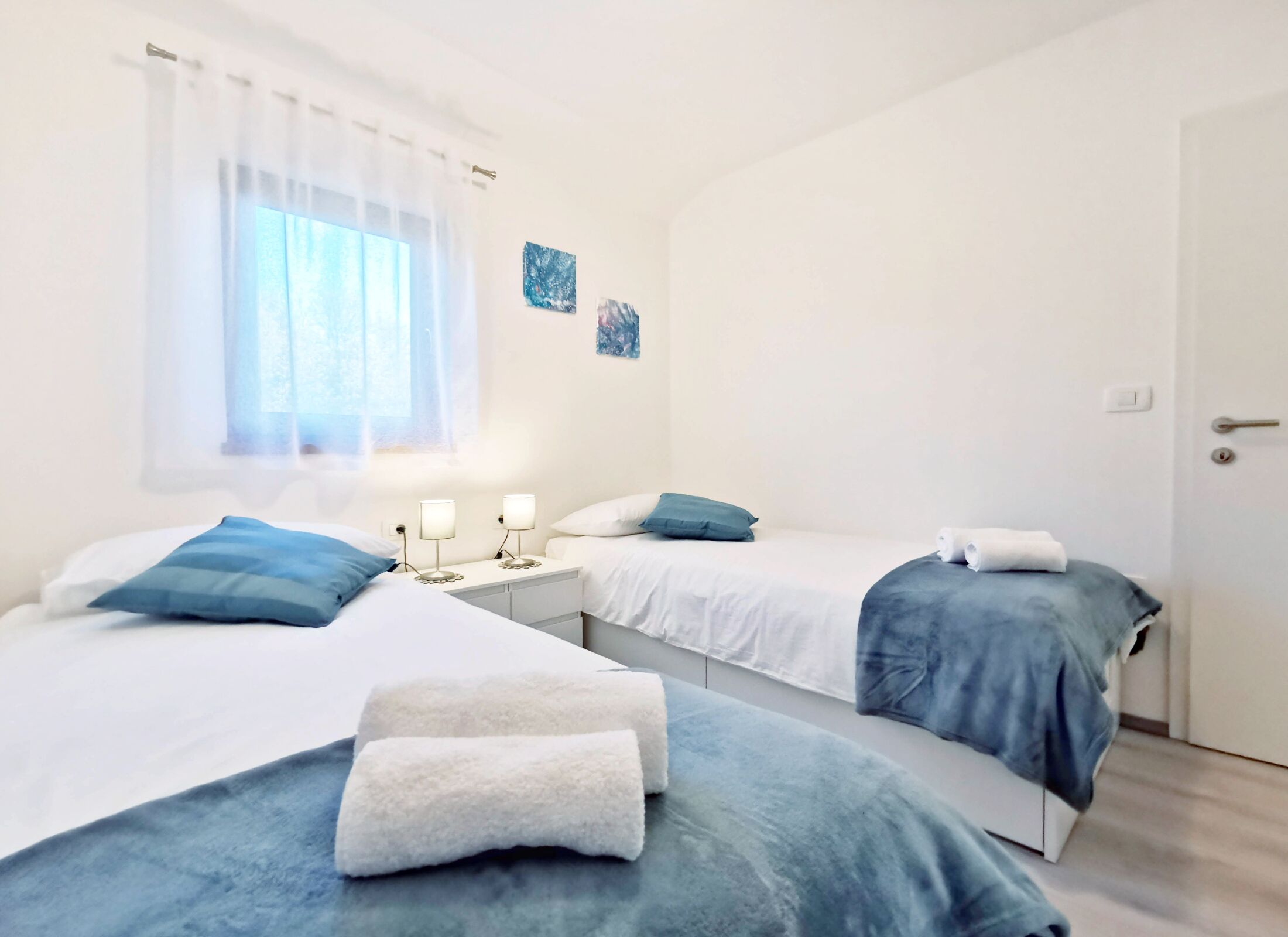 A hotel room with two beds, each with a blue blanket and white pillows. There are two bedside tables with lamps, a window with sheer curtains, and a door.