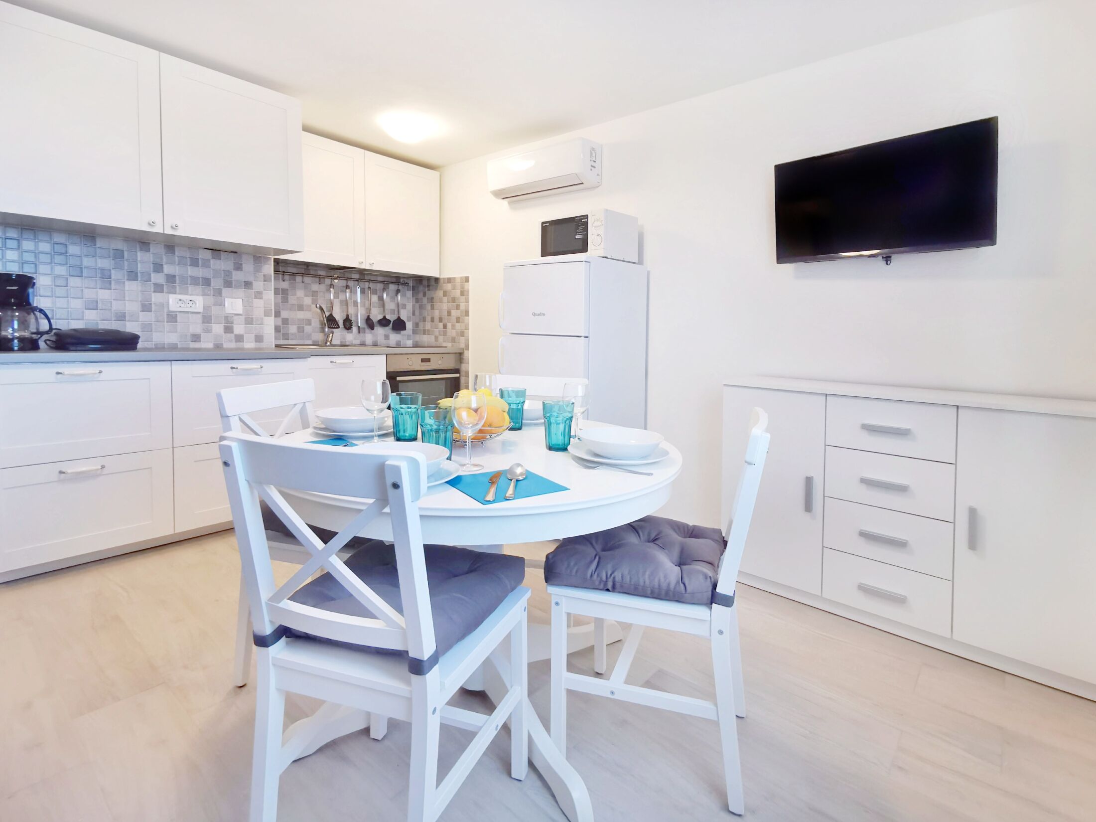A modern kitchen with white cabinets, a tiled backsplash, and a flat-screen TV mounted on the wall.