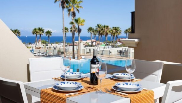 A dining table set with plates, wine glasses, and a bottle, overlooking a pool and palm trees with a view of the ocean. - El Faro by Interhome (Palm- Mar)