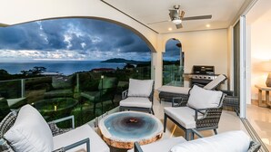 A balcony with a glass enclosure, white cushioned chairs, a round table with a central fire feature, and a grill. - La Vista #B10, amazing Views - Brand New 3 bd Condo overlooking Tamarindo Bay Vista B-10 (Tamarindo)