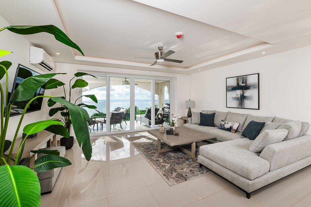 A modern living room with a grey sofa, a coffee table, and large windows offering a view of the outdoors.