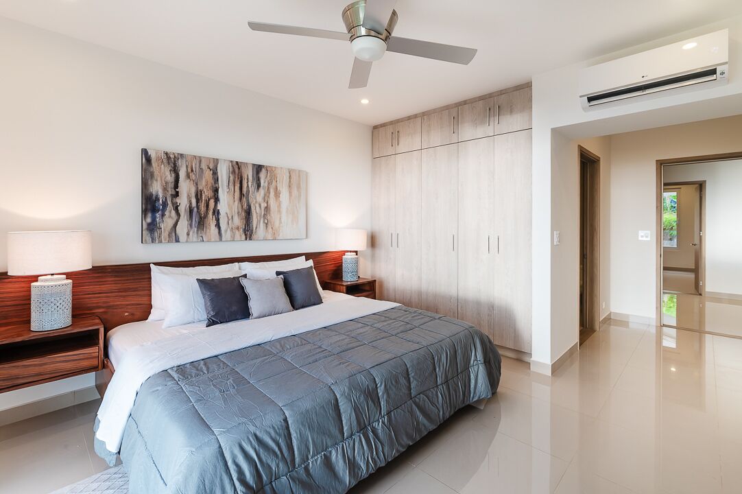Un dormitorio moderno con una cama grande, cabecera de madera, dos mesitas de noche con lámparas, un ventilador de techo y un cuadro abstracto de gran tamaño en la pared.