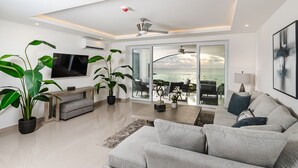 A modern living room with a large flat-screen TV, a grey sofa, and a bench. There are several potted plants and a ceiling fan. The room has a large sliding glass door leading to a balcony with a view of the ocean. - La Vista #B10, amazing Views - Brand New 3 bd Condo overlooking Tamarindo Bay Vista B-10 (Tamarindo)