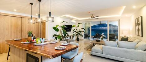 A modern kitchen with a central island, dining area, and a living room with a sofa and a TV.