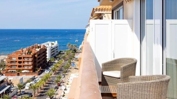 A balcony with wicker furniture, a view of the sea, and a building with balconies in the background.