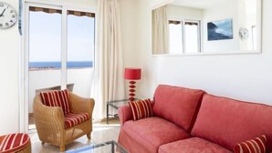 A living room with a red sofa, wicker armchair, and a glass coffee table. There is a large window with a view of the sea.