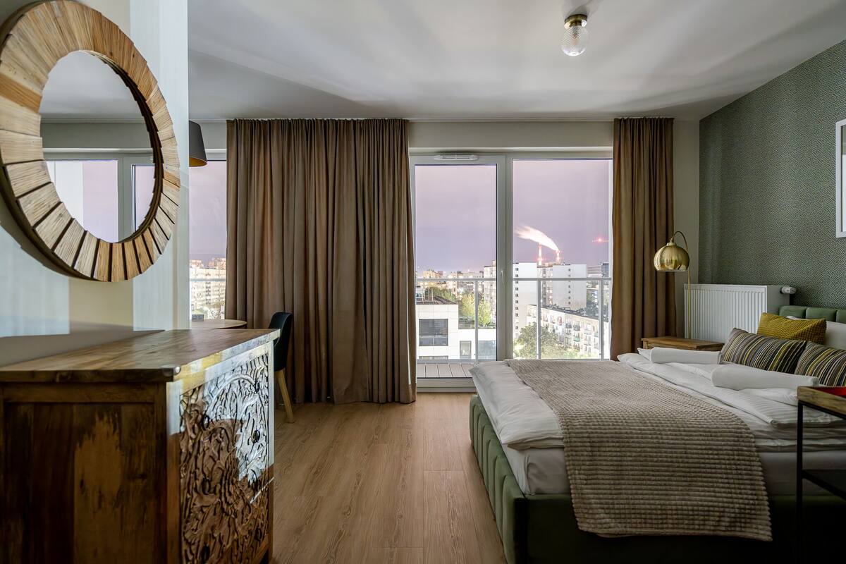 A modern hotel room with a large bed, a wooden desk, a round mirror, and a view of the cityscape through large windows.
