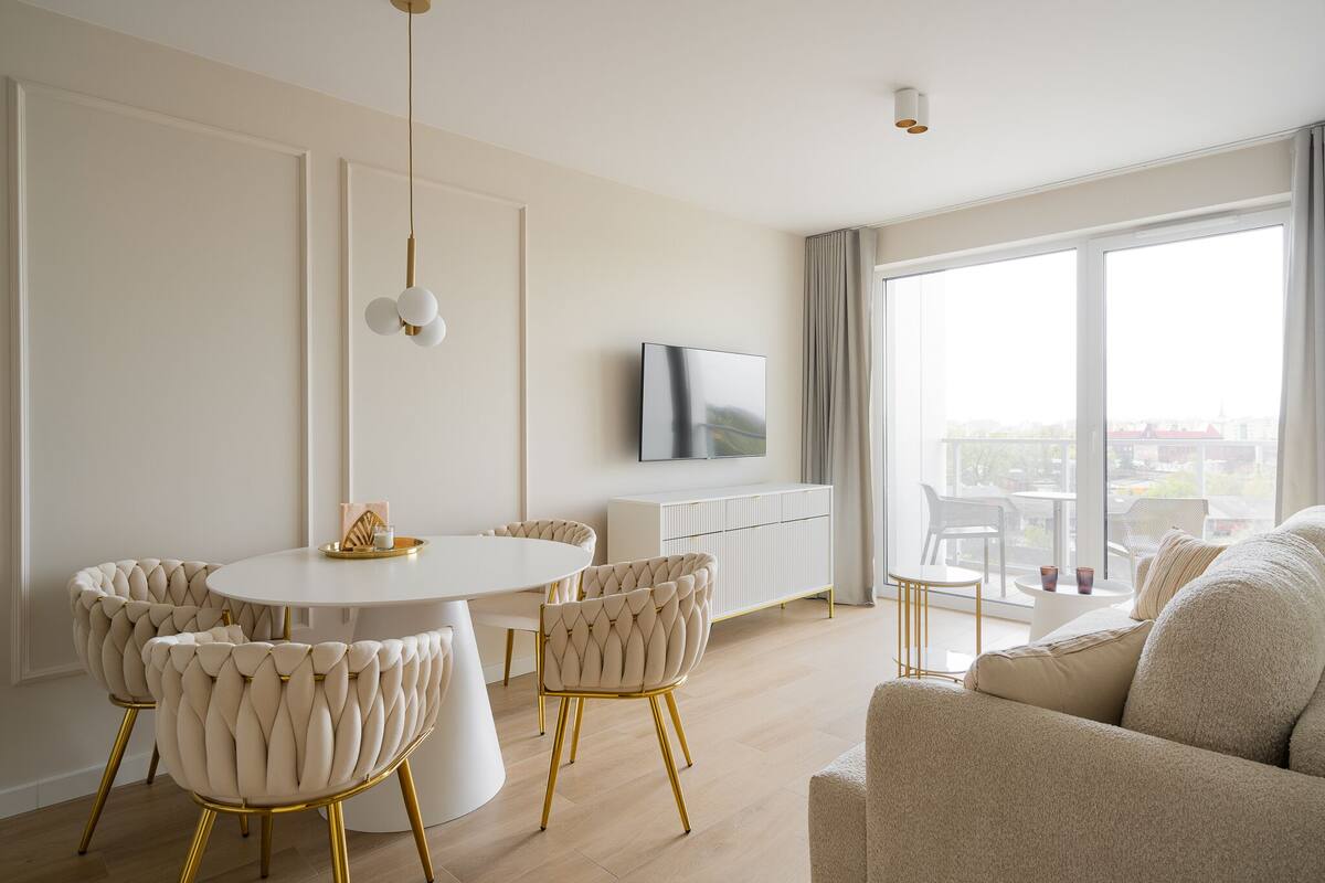 A modern living room with a round dining table, tufted chairs, a sofa, a TV, and a large window with a city view.